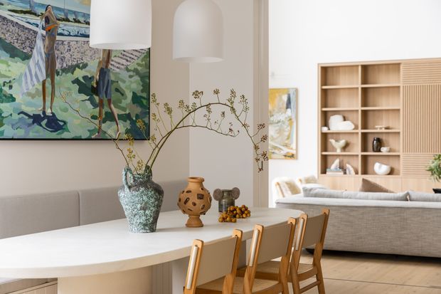 A bright, refined open-plan dining and living area. A large white dining table with timber chairs occupies the foreground, with a neutral upholstered sofa and built-in timber shelving in the living area beyond. The room features arched wall niches, sculptural pendant lights, pale timber flooring, neutral walls, and colourful artwork with organic floral branches as decor.