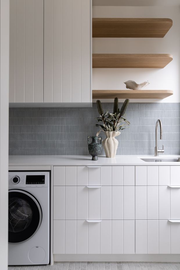 A bright, compact utility space combining a laundry area with a kitchenette-style sink and extensive white built-in cabinetry. Vertical white wall panelling and cabinet fronts contrast with a fine grey vertical-tile splashback, while three floating timber shelves add warmth above the benchtop. A front-loading washing machine, undermount sink with brushed metal tapware, decorative vases and subtle styling are visible.