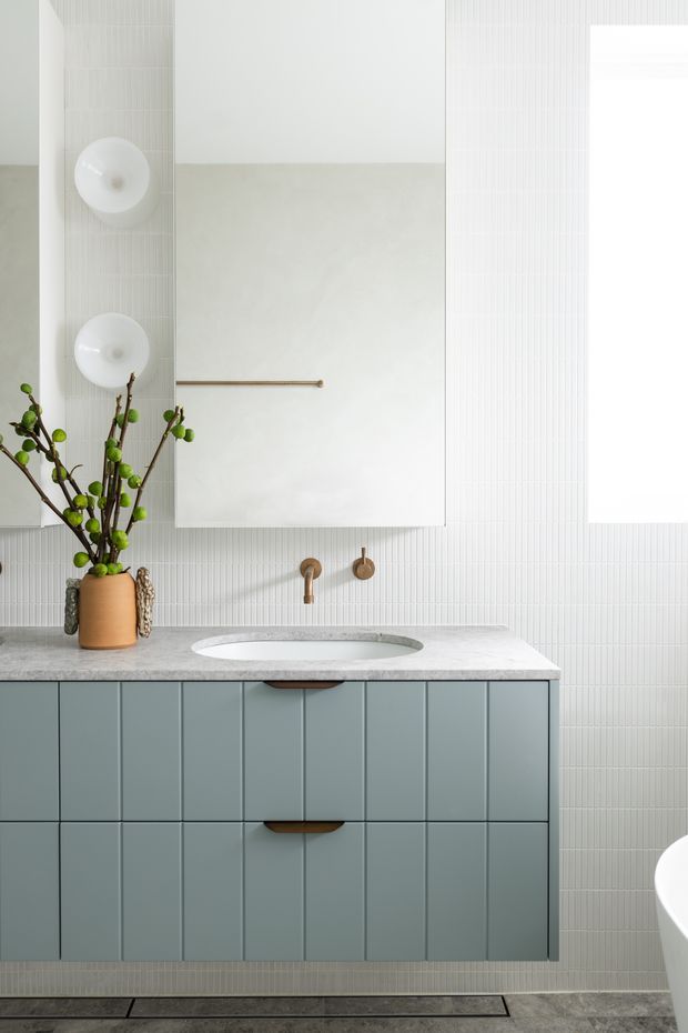 A bright, refined contemporary bathroom vanity with a floating blue-grey tiled cabinet, white stone benchtop and integrated oval basin. Vertical white wall tiles, a large frameless mirror, brass wall-mounted tapware and a slim brass towel rail create a clean, softly textured look. A freestanding bathtub is partially visible at right, with a decorative branch arrangement adding warmth.