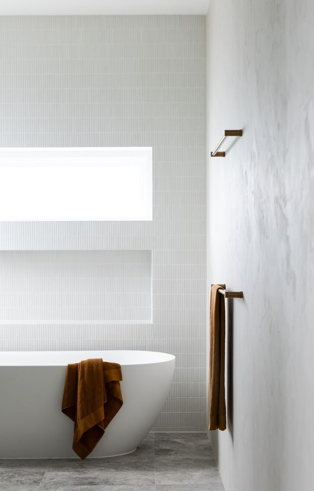 A serene contemporary bathroom featuring a freestanding white bathtub, pale vertically ribbed wall tiles, a large frosted or overexposed window, grey stone-look floor tiles, and warm bronze towel rails with a brown towel. The space has a clean, minimalist composition and soft natural light.