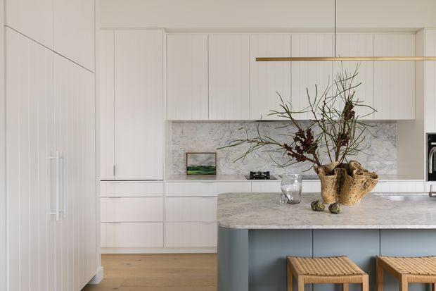 A bright, refined contemporary kitchen with full-height white paneled cabinetry, a pale stone splashback and island benchtop, and a muted blue-grey island base. Two woven timber stools sit at the island, while a linear pendant light and sculptural floral arrangement add understated decorative detail. Timber flooring and soft neutral tones create a calm, airy atmosphere.