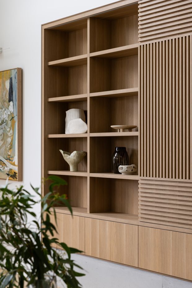 Close-up interior view of a custom built-in timber shelving and cabinetry unit. Open display niches with horizontal shelves sit beside vertically slatted and horizontally ribbed cabinet fronts, all in a warm light-to-mid timber finish. Neutral white walls and flooring frame the joinery, with a few ceramic vessels, artwork, and a partially visible plant adding restrained decorative detail.