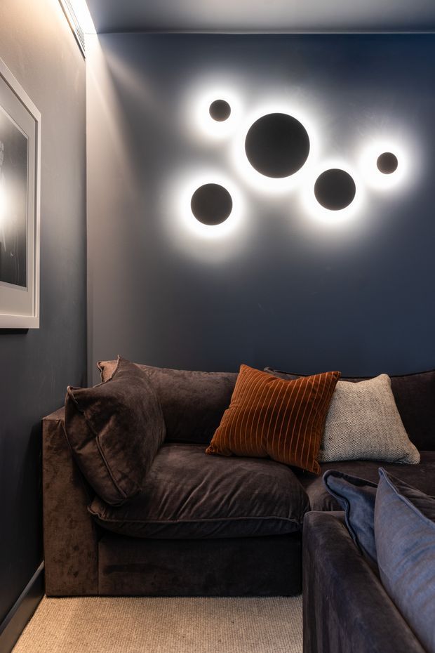 A compact, moody living room or media lounge with deep navy walls, dark velvet sectional seating, warm rust and neutral cushions, and a beige carpet. A cluster of round black wall lights creates a dramatic halo effect above the sofa, while framed wall art is partially visible at left.