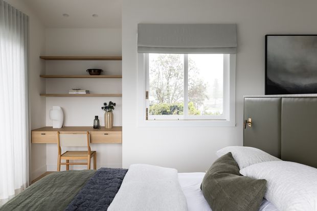 Bright, softly styled bedroom with a built-in timber desk and open shelving, upholstered bedhead, layered white and olive bedding, a large window with a Roman blind, and sheer curtains. The room has a calm, contemporary and neutral appearance with integrated storage and workspace.