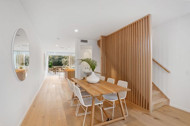 Bright open-plan dining area with a light timber dining table and white chairs, positioned beside a vertical timber slat privacy screen and staircase. Pale timber flooring, white walls, recessed lighting, and a large round wall mirror create an airy contemporary interior. Glazing at the rear opens toward an outdoor area.