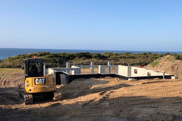 A coastal construction site showing a partially built concrete building foundation and walls, with a CAT excavator in the foreground. Low scrub-covered dunes and the ocean are visible beyond the site under a clear blue sky.
