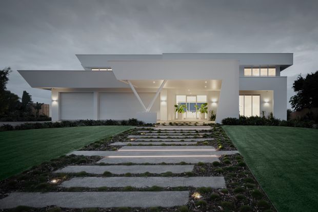 A dramatic dusk exterior view of a sleek, low-profile modern residence. The white geometric facade features broad flat roof planes, large glazed openings, recessed entry lighting, integrated garage doors, and a prominent diagonal structural support. A landscaped front lawn and illuminated stepping-stone path lead toward the entrance.