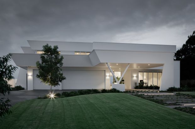 A contemporary white two-storey residence viewed from the landscaped front yard at dusk. The angular flat-roofed facade features broad projecting forms, recessed warm lighting, a covered entry or carport zone, large glazed openings, and a prominent white V-shaped structural support. A manicured lawn, low planting, and stepping-stone driveway occupy the foreground beneath an overcast sky.
