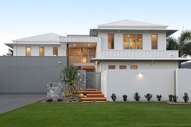 Contemporary multi-level residential facade with light grey horizontal cladding, broad low-pitched metal roofs, timber louvre screens, and a sheltered entry. The property features a tall privacy fence and gate, landscaped lawn, stone-clad entry pillar, stepped timber entry stairs, and warm exterior lighting.