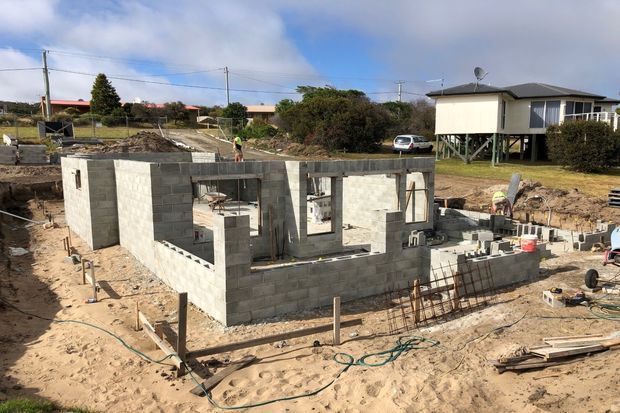 A single-storey residential building under construction on a sandy site. The partially built structure has grey concrete block walls, open window and door openings, exposed reinforcement steel, and unfinished foundations. Nearby detached homes and utility poles are visible in the background.