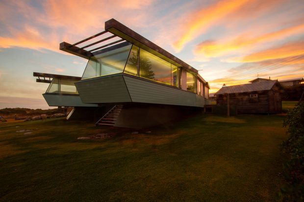 Exterior view of a distinctive elevated modern dwelling at sunset, with a long angular form, extensive floor-to-ceiling glazing, projecting roof structures, and dark horizontal cladding. The house is raised above the grassy site and sits beside a rustic timber outbuilding in a rural or coastal-looking setting.
