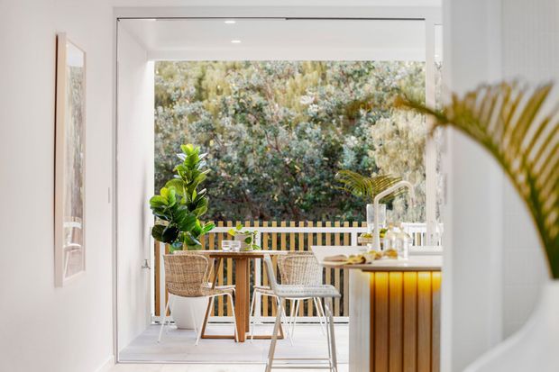 Bright contemporary kitchen and dining space opening directly onto a small outdoor terrace or balcony. The room features white walls and cabinetry, a timber dining table, woven dining chairs, a compact island or breakfast counter with warm timber fluting and a light benchtop, and large open glazing framing dense greenery outside.