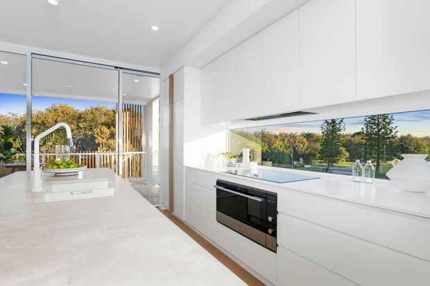Bright contemporary white kitchen with a large pale stone island, integrated white cabinetry, built-in black oven, stainless steel gooseneck tap, and expansive glazing overlooking a landscaped suburban setting. The space is clean-lined and filled with natural light.