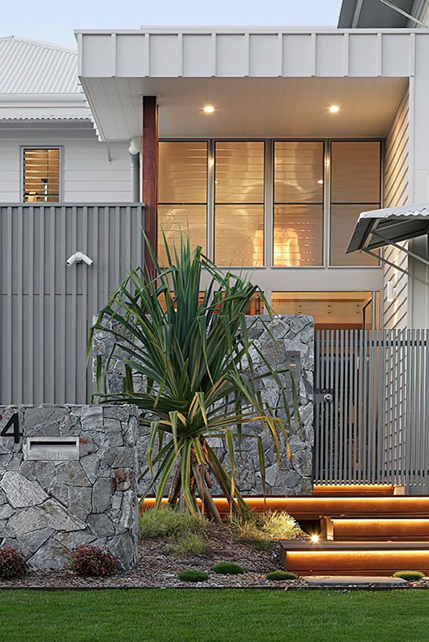 A contemporary multi-level residential facade viewed from the landscaped front approach. The house combines pale horizontal cladding, grey metal privacy screens and roofing, large timber-toned glazed windows, dark timber accents, and a rugged natural-stone retaining wall. Illuminated timber-edged entry steps lead up through a planted front garden, with a visible house number and garage structure at right.