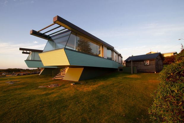 Exterior view of a distinctive elevated modern house with a long, angular form, extensive glazing, projecting roof beams, and dark horizontal cladding. The building is raised above the grass on a sculptural structural base with an external stair, and includes a small separate dark timber outbuilding. The setting appears open and suburban or semi-rural, photographed in warm late-day light.