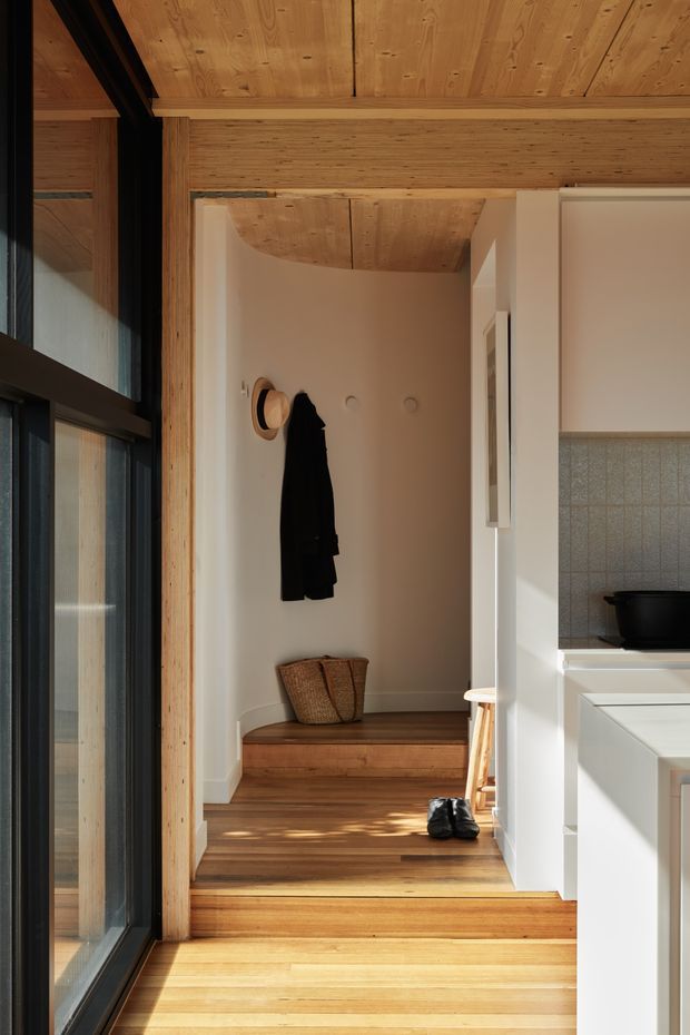 Warm, compact residential entryway opening directly into a kitchen. The space features extensive natural timber lining on the ceiling, walls and stepped flooring, black-framed glazing at left, white cabinetry at right, and a small recessed coat and shoe storage area with hooks, a hat and baskets. Strong sunlight creates a calm, refined atmosphere.