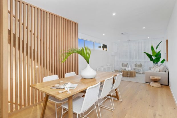 Bright open-plan dining and living area with a light timber dining table and white wire-frame chairs. A vertical timber slat screen partially separates the dining zone from the lounge, which features pale upholstered seating, a round timber coffee table, sheer white curtains, indoor greenery, and light timber flooring. Recessed ceiling lights and high clerestory-style windows contribute to an airy contemporary interior.