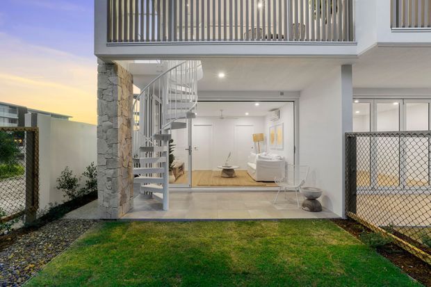 Contemporary two-storey residential facade viewed from a small landscaped courtyard or front garden. The open ground-level living area features white walls, light timber flooring, a white sofa and coffee table, and extensive glazing. A striking white spiral staircase rises beside a rough natural-stone-clad column, while grey metal vertical balustrades define the upper-level balcony. Chain-link fencing and lawn frame the exterior at dusk.