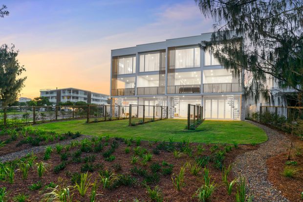 Exterior architectural photograph of a contemporary multi-storey residential building at dusk. The house has a rectilinear form, extensive floor-to-ceiling glazing, light-coloured rendered or panelled walls, dark window frames, balconies, and a landscaped foreground with lawn, garden beds, fencing, and a gravel path.