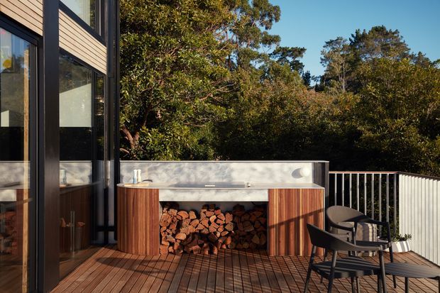 A contemporary outdoor deck beside a dark-framed, timber-clad home, featuring a built-in outdoor fireplace or firewood storage bench with a pale rendered top and vertical timber sides. A small black outdoor dining setting sits on the timber decking, with dense mature planting beyond the railing.