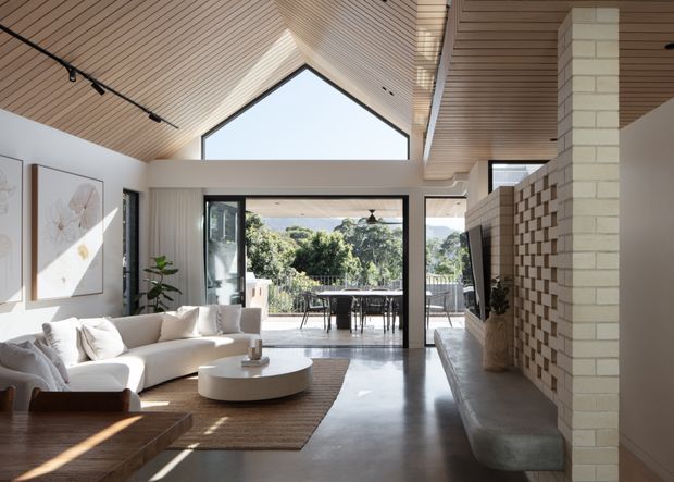 Bright contemporary living room with a dramatic vaulted timber-lined ceiling, exposed triangular gable glazing, polished concrete flooring, and large black-framed doors opening to an outdoor dining terrace. The room includes a light sectional sofa, round coffee table, built-in concrete bench seating, a white brick fireplace or feature wall, and an open connection to a leafy exterior.