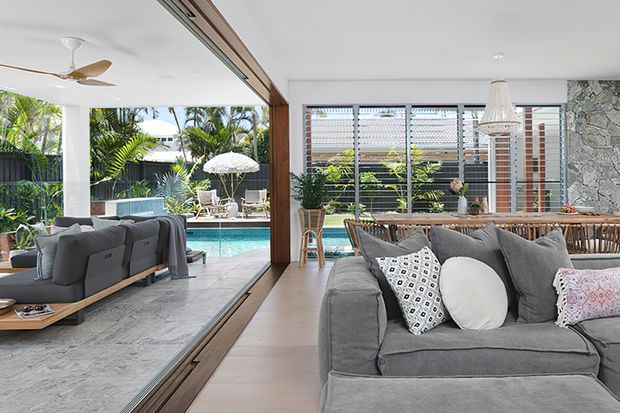 Open-plan contemporary living area with a large grey sectional sofa in the foreground, connected through wide timber-framed glazed doors to a covered outdoor entertaining space and swimming pool. The room features light timber flooring, white walls, extensive horizontal louvred glazing, a stone feature wall, a woven pendant light, and a tropical landscaped outlook. A second seating area and ceiling fan are visible in the outdoor zone.