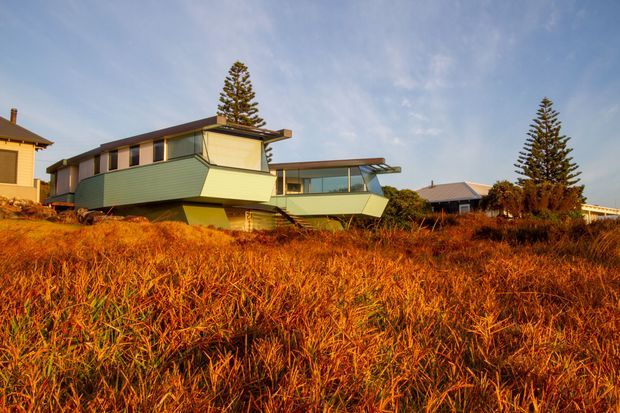 Exterior view of a distinctive mid-century-inspired coastal residence set behind tall golden dune grass. The house has a low, angular form with broad projecting roof planes, horizontal strip windows, pale green cladding, and dark timber or metal framing. Neighbouring homes and tall conifer trees are visible in the background under a clear blue sky.