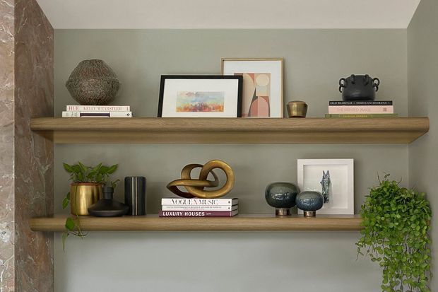 Close-up view of two built-in timber display shelves set against a muted sage-grey wall, styled with framed artwork, books, ceramics, decorative objects, and trailing plants. A stone or marble-clad vertical wall edge is visible at the left. The composition has warm natural materials and carefully arranged contemporary decor.