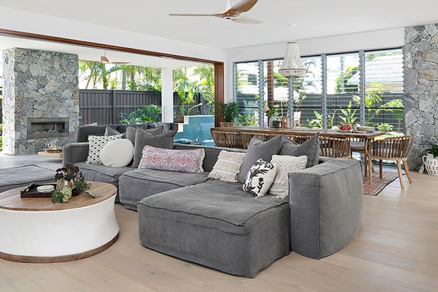 Bright open-plan living and dining room with a large grey sectional sofa, round white coffee table, timber dining table and chairs, extensive louvred glazing, pale timber flooring, and a stone-clad fireplace. Indoor plants and woven furnishings add a relaxed coastal feel, with an outdoor garden visible beyond the glazing.