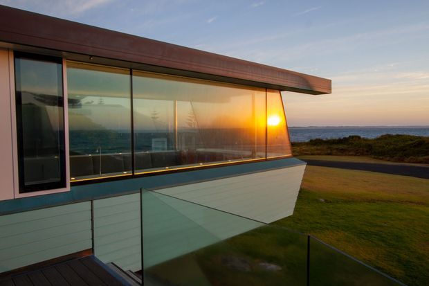 Exterior architectural photograph of a sleek, low-profile coastal building at sunset. The composition highlights a projecting flat roof, extensive floor-to-ceiling glazing with dark frames, pale horizontal cladding, and a glass balustrade above a deck or terrace. The ocean horizon and shoreline are visible beyond the building, reinforcing the waterfront coastal setting.