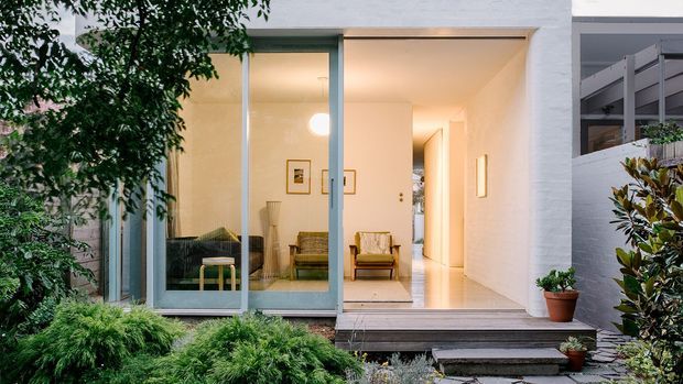 A contemporary house exterior viewed from a landscaped garden, with a white rendered facade and broad floor-to-ceiling glazed sliding doors opening into a bright, sparsely furnished living room. The interior features pale walls and flooring, timber-framed lounge chairs, framed artwork, and a warm pendant light. A timber deck and steps connect the living space to the garden.