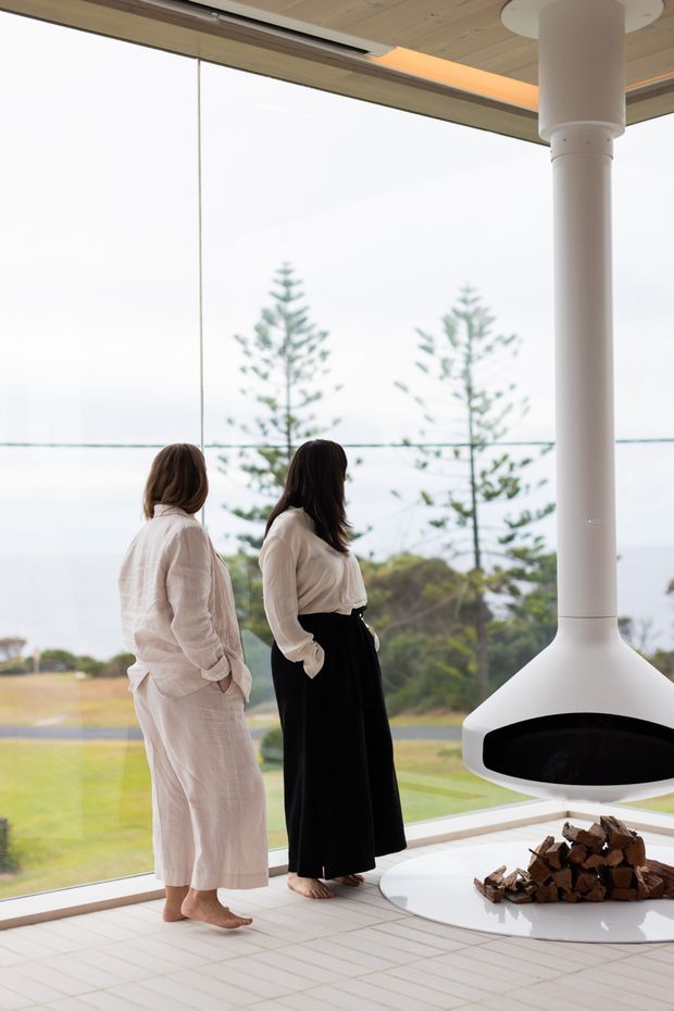 A bright, refined interior with floor-to-ceiling glazing overlooking a landscaped coastal setting. Two people stand beside a large suspended white fireplace with a broad cylindrical flue and a circular hearth containing stacked firewood. The room has pale timber ceiling lining, light tiled flooring, white structural columns, and expansive views through the glass.