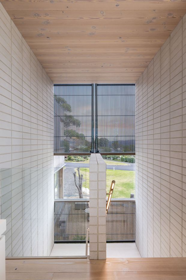 A bright, vertically composed interior view down a stairwell or landing. Pale rectangular masonry tiles line both side walls, while the ceiling and stair treads are clad in warm natural timber boards. A central tiled structural wall divides the stair opening, with slim vertical dark metal screening across large windows revealing a landscaped suburban exterior. A timber handrail and stair elements are visible, creating a clean, modern architectural composition with strong daylight and layered views.