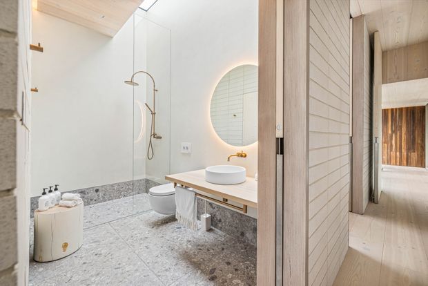 A bright contemporary bathroom viewed from an adjoining hallway, featuring a walk-in glass shower with brushed brass fixtures, a wall-mounted toilet, a floating timber vanity with a white vessel basin, and a round backlit mirror. The bathroom has terrazzo-like grey flooring and a matching low wall finish, white walls, pale timber ceilings and joinery, and a softly illuminated, refined natural palette. Pale timber-lined hallway elements and a glimpse of another timber-clad space are visible beyond.