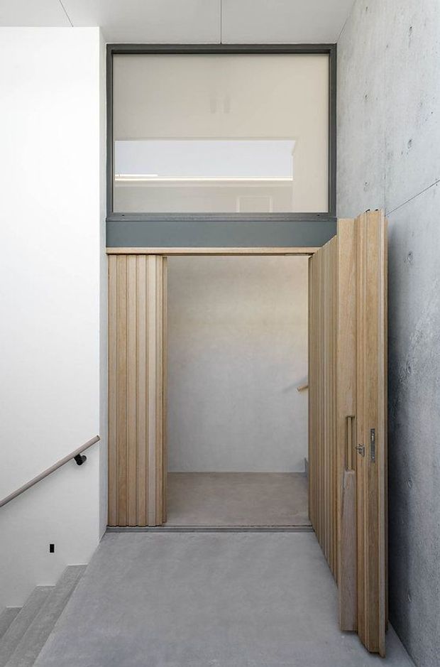 A minimalist concrete stair landing and hallway with a partially open vertical timber-slat door. The space features smooth grey concrete flooring and walls, a dark metal-framed internal window or transom above the doorway, white plaster walls, and a simple metal handrail. The view looks toward a small adjoining room through the open door.
