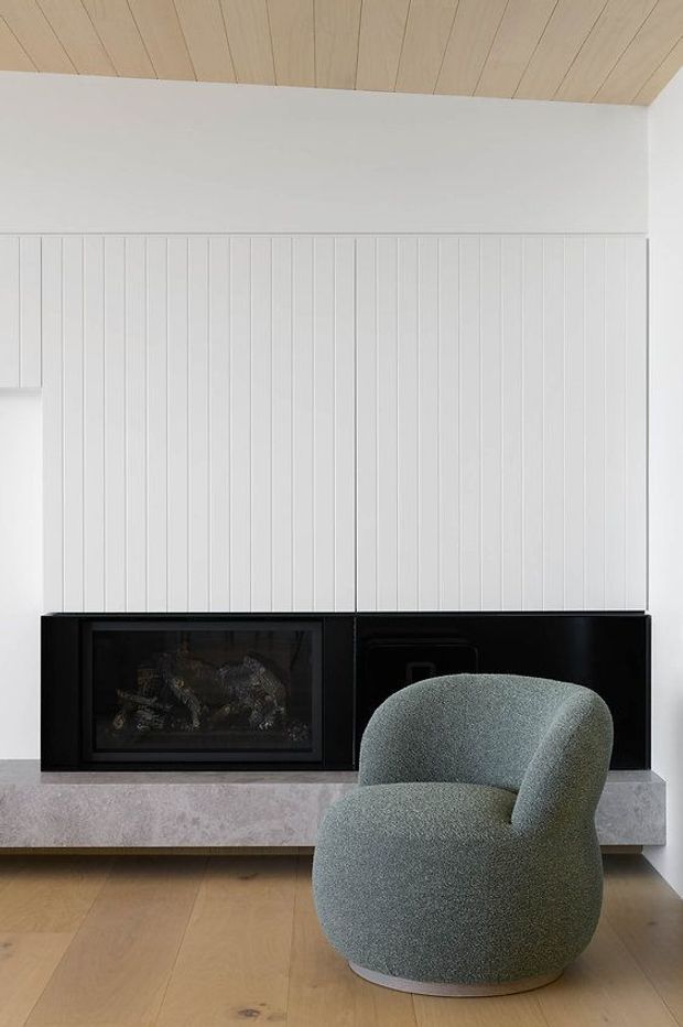 A refined living room featuring a black built-in fireplace set into a pale stone hearth, against a white vertically panelled feature wall. The room has a light timber-lined ceiling and matching timber floor, with a sculptural muted green upholstered lounge chair in the foreground. The composition is minimalist and contemporary with warm natural materials and strong black-and-white contrast.