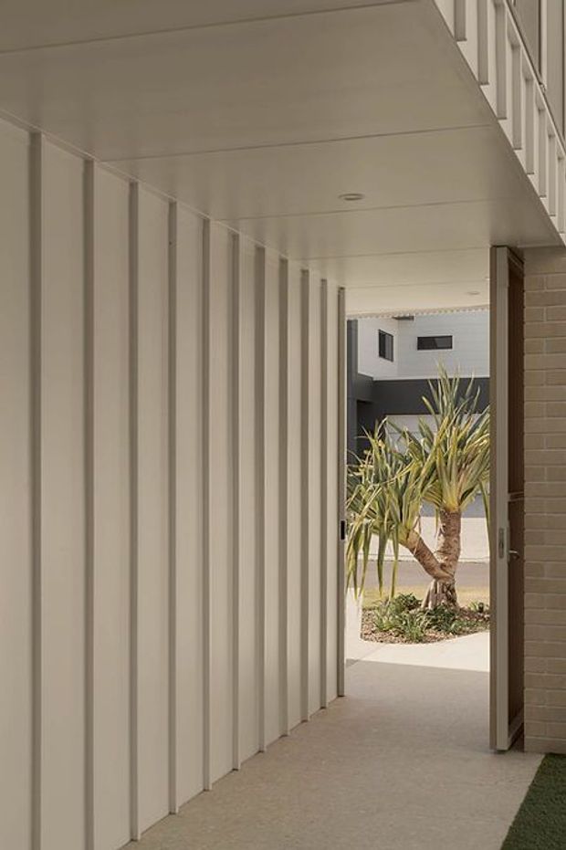 A covered exterior walkway or breezeway with a smooth pale concrete floor, white vertical wall panels, a white soffit with recessed lighting, and a warm brick wall at the entrance. The passage opens onto a landscaped courtyard with a palm-like plant and a contemporary building visible beyond.