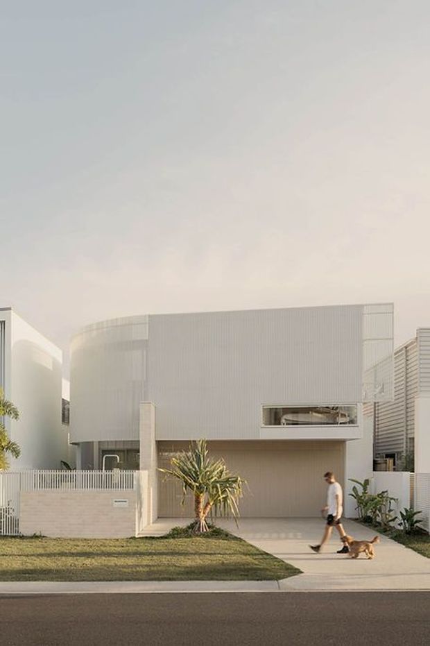 Street-facing view of a contemporary multi-level residence with a softly curved upper facade, pale vertical ribbed cladding, crisp white rendered forms, a recessed garage, and a narrow horizontal window. The front garden has lawn, a sculptural yucca-like planting and low masonry fencing, with a pedestrian and dog on the footpath.