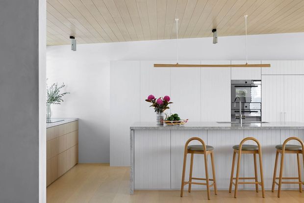 Bright contemporary kitchen with a long island featuring vertically ribbed cabinetry, a grey stone benchtop, and three timber bar stools. Full-height white cabinetry houses integrated ovens, while a timber-lined ceiling, light timber flooring, pale timber side cabinetry, and linear pendant lighting create a warm, minimal interior.