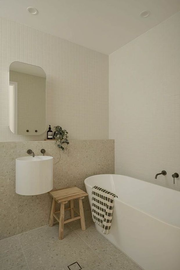 A warm, minimalist bathroom with a freestanding white vessel basin, wall-mounted dark metal taps, a rounded rectangular mirror, and a white bathtub. Pale terrazzo-style surfaces cover the floor and lower wall, while vertically textured pale wall tiles add subtle detail. A small timber stool and woven towel provide natural accents.