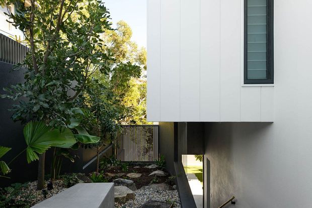 A modern residential exterior viewed along a narrow landscaped passage. The image shows a white vertically panelled upper volume cantilevered over a recessed ground-level walkway, with a slim dark-framed window, dark vertical screening, concrete surfaces, gravel and lush planting. The composition emphasizes clean lines, layered privacy screens and a sheltered connection through the building.