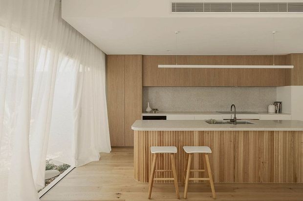 Bright contemporary kitchen with a long timber island featuring rounded ends and vertical ribbed detailing, pale stone benchtops, matching timber cabinetry, and two light timber stools. Sheer white curtains line the full-height glazing at left, while a subtle textured grey splashback and integrated ceiling ventilation complete the calm, warm-toned interior.