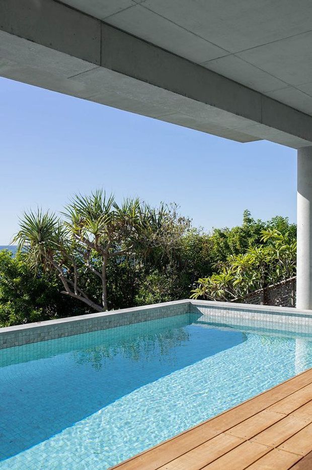 A bright outdoor swimming pool beneath a broad concrete roof overhang, viewed from a timber deck. The pool has pale blue mosaic tile lining and a raised tiled edge, with lush tropical planting and clear sky beyond. The image emphasizes the pool, shaded architectural structure, and warm timber decking.