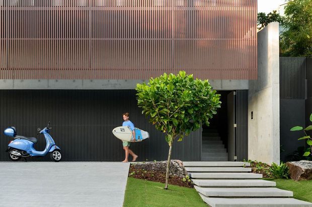 Exterior view of a contemporary multi-level residence with a vertically screened upper facade in warm brown tones, dark ribbed garage doors, exposed concrete walls, and a stepped concrete entry path. A landscaped front garden, scooter, and person carrying a surfboard are visible.