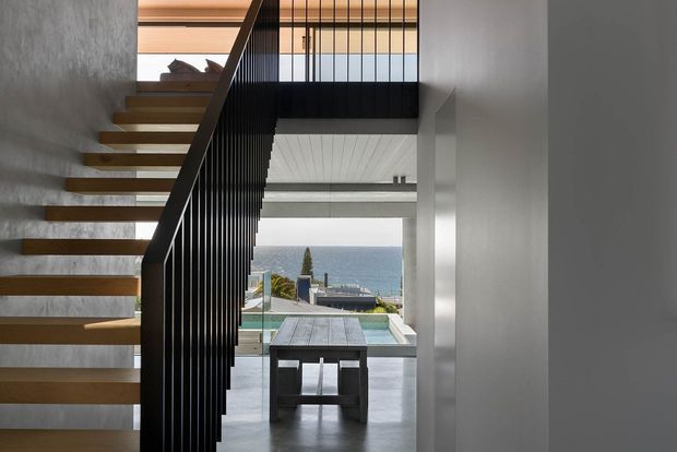 A modern interior stairway with floating timber treads, black vertical metal balustrades, and concrete-toned walls. The stair rises beside a double-height opening toward an upper landing, while a lower-level dining or living area and a swimming pool are visible beyond, with a coastal ocean view in the distance. The space is clean-lined, open, and architecturally minimalist.
