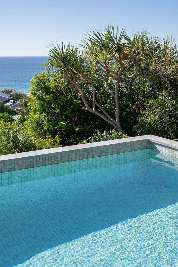 A sunlit outdoor swimming pool with turquoise mosaic tile lining and pale concrete coping, surrounded by lush tropical landscaping. The pool overlooks a blue ocean visible beyond the trees, creating a coastal, elevated setting.