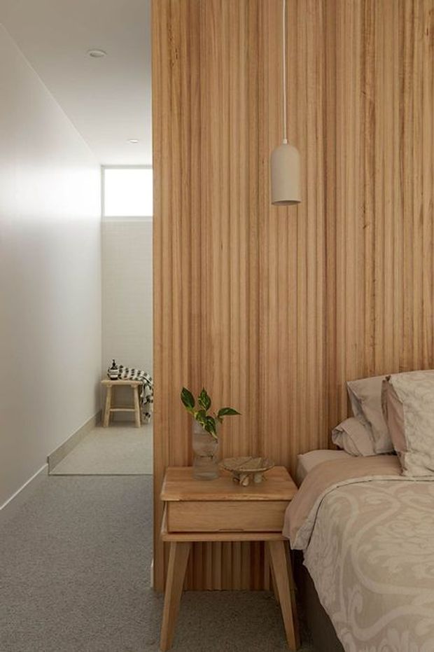 A warm, minimalist bedroom with a timber-lined feature wall, matching timber bedside table, pendant light, and a neatly styled bed in soft neutral bedding. A compact tiled bathroom or dressing area is visible beyond the open passage, with a small stool and high window. The room has a calm, natural material palette and soft daylight.