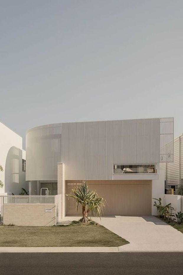 A contemporary multi-storey residential facade viewed from the street. The house features a curved upper-level form clad in fine vertical light-grey slats, a recessed garage, narrow horizontal window, pale masonry boundary wall, and a simple front lawn with drought-tolerant planting. The composition is clean and minimalist with soft neutral tones.