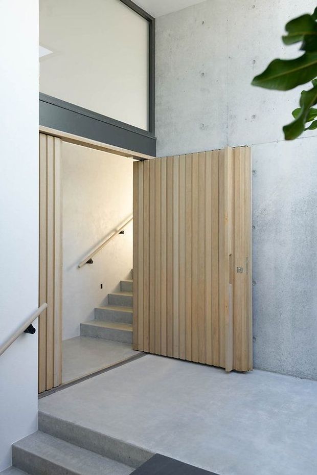 A minimalist interior stair entry featuring a tall pivot door clad in closely spaced light timber slats, exposed smooth concrete walls and flooring, and a black-framed upper glazed opening. Concrete stairs lead through the timber doorway, with simple metal handrails and a clean, high-ceilinged composition.