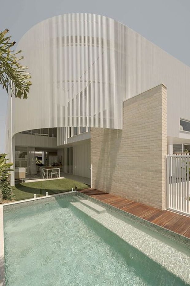 Exterior architectural photograph of a contemporary multi-level residence centred on a narrow swimming pool and courtyard. The building features a sculptural curved white vertical-slat facade, pale masonry walls, large glazed openings, timber decking, and a white vertical-bar gate. An outdoor dining area and interior living spaces are visible beneath the cantilevered upper level.